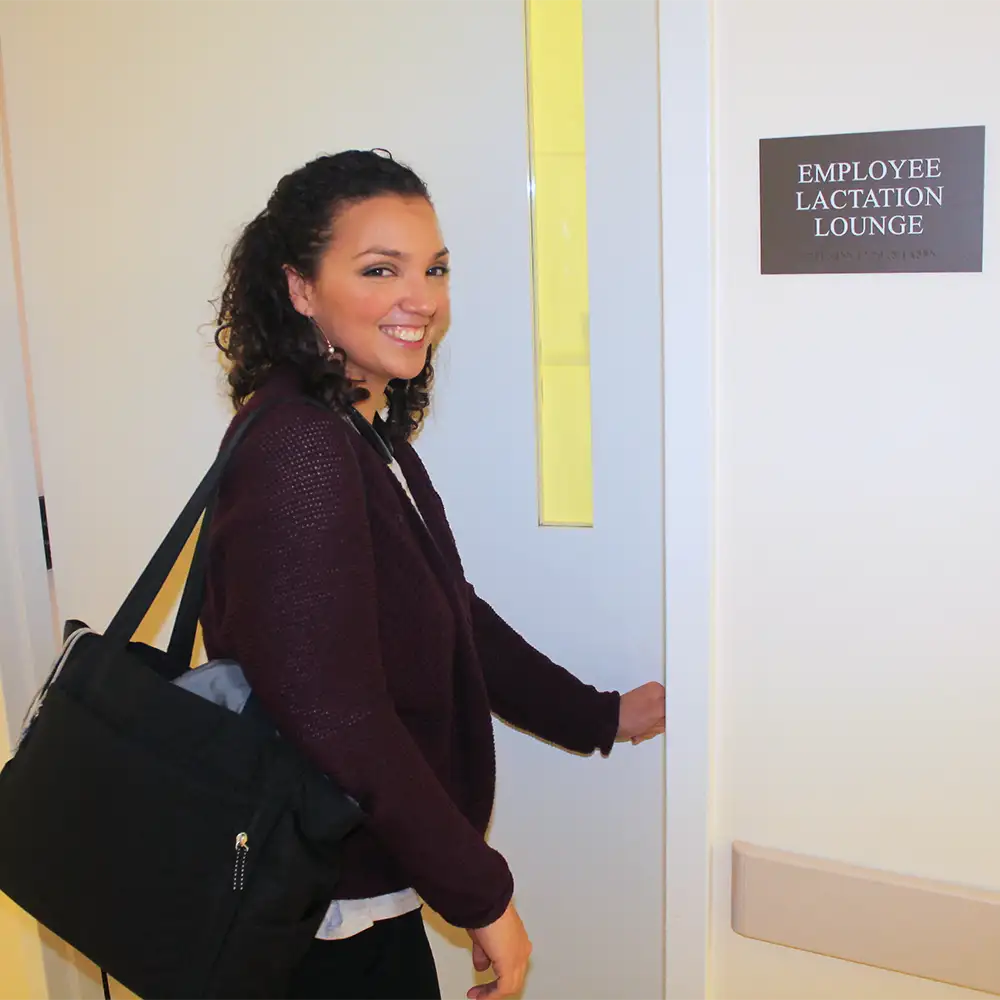 A woman with a bag on her shoulder smiling and walking into an employee lactation lounge
