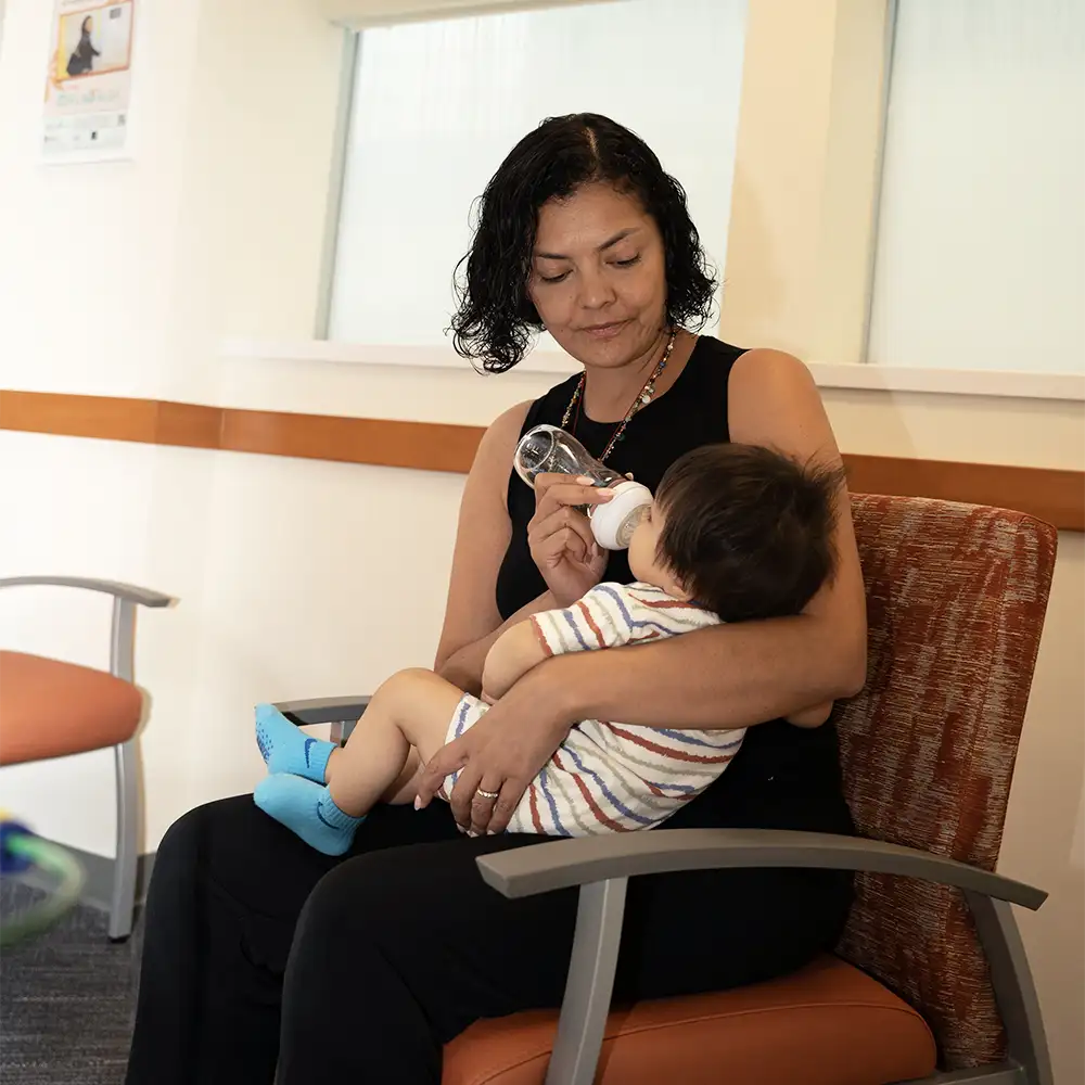 A woman with dark curly hair sitting in a chair feeding her baby with a bottle 