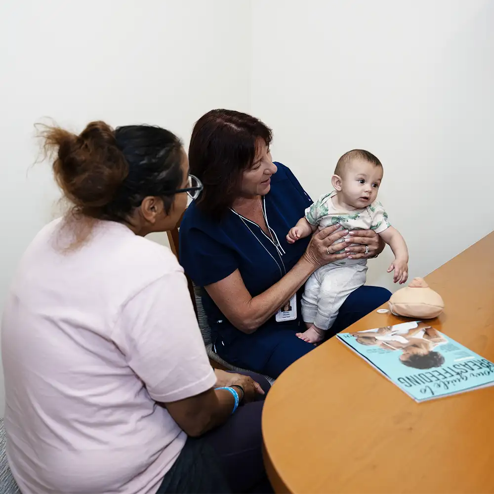 A female physician holding an infant while his mother is right next to them with a Breastfeeding magazine on the table infront of them
