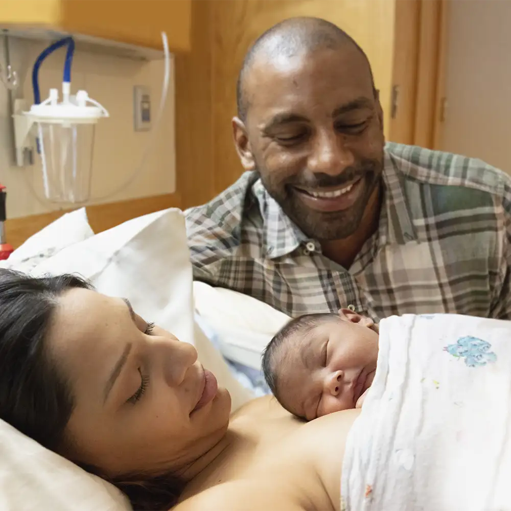 A mom sleeping in a hospital bed with her newborn on her chest while the father looks down on them smiling